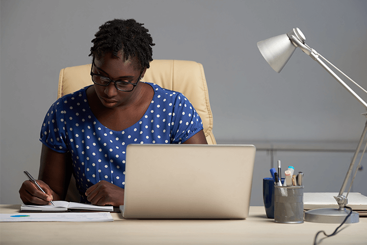 Mulher negra sentada à mesa escrevendo em um caderno com um notebook próximo de si exercendo a profissão de planejador financeiro