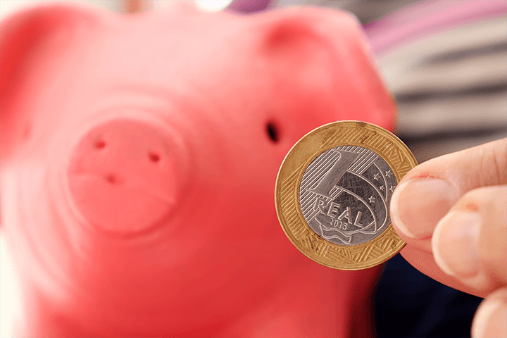 Cofrinho em formato de porco rosa desfocado ao fundo e dedos segurando uma moeda de 1 real. Selic e Inflação. 